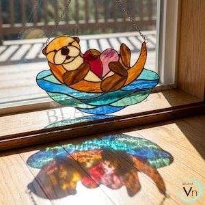 Pode incluir: Apanhador de sol de vidro manchado com uma lontra. A lontra é em tons de castanho e bege, segurando dois corações vermelhos e rosa. Flutua sobre água azul e verde. O apanhador de sol é pendurado por uma corrente.
