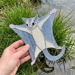 May include: Stained glass sugar glider with outstretched wings, in shades of blue, white, and pink. The glass art piece is held against a backdrop of green grass and water, showcasing its intricate design and craftsmanship.