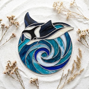 Puede incluir: Un atrapasoles circular de vidrieras que muestra una orca blanca y negra nadando a través de olas oceánicas en tonos azul profundo, turquesa y blanco. Las piezas de vidrio están unidas por finas líneas de soldadura de metal plateado.