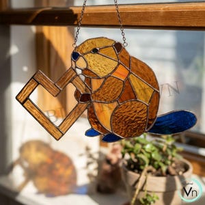 May include: Stained glass beaver suncatcher with a wooden heart-shaped frame. The beaver is made of brown, orange, and yellow glass, with a blue tail and eyes. It hangs from a chain, perfect for a window or wall decoration.
