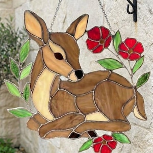 Könnte beinhalten: Glasmalerei-Hirsch mit roten Blumen und grünen Blättern. Der Hirsch ist braun und cremefarben, mit einem schwarzen Auge und einer schwarzen Nase. Die Blumen sind rot mit grünen Blättern. Das Kunstwerk hängt an einer schwarzen Metallhalterung.