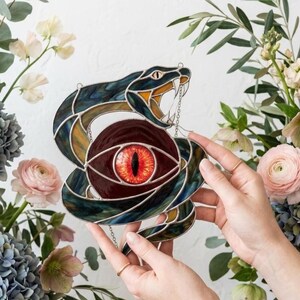 Puede incluir: Un atrapasoles de vidrieras que muestra una serpiente enroscada con escamas de vidrio iridiscente azul oscuro y verde, rodeando una pieza central de vidrio circular con un ojo naranja y rojo detallado y realista con una pupila vertical negra.