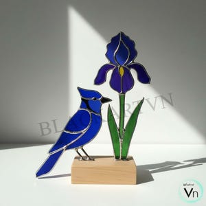 May include: Stained glass art piece featuring a blue jay and a purple iris flower. The bird is perched on a wooden base next to the flower. The iris has green leaves and a yellow center. The artwork is signed with the text "blackart Vn".