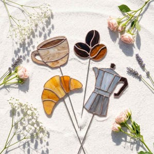 May include: Four stained glass plant stakes featuring a coffee cup in marbled beige, a golden yellow croissant, a dark brown coffee bean cluster, and a light blue moka pot. Each piece is crafted with metal soldering and attached to a thin metal rod.