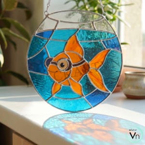 Könnte beinhalten: Buntglas-Suncatcher mit einem orangefarbenen Goldfisch in einer runden Schüssel. Der Fisch hat schwarze Details, vor einem blau-türkisfarbenen Hintergrund. Die Schüssel ist klar mit einer silbernen Kette zum Aufhängen.