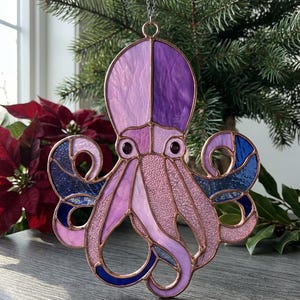 Könnte beinhalten: Glasmalerei-Oktopus-Ornament in Lila-, Rosa- und Blautönen. Der Oktopus hat ein detailliertes Design mit kupferfarbenen Umrissen und hängt an einer silbernen Kette. Das Ornament wird vor einem Weihnachtsbaum präsentiert.