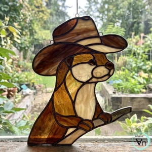 Pode incluir: Um apanhador de sol em vitral representando uma lontra usando um chapéu de cowboy de abas largas. A peça é feita de vários tons de vidro castanho e creme, unidos por linhas de solda de metal escuro, e possui um laço para pendurar no topo.
