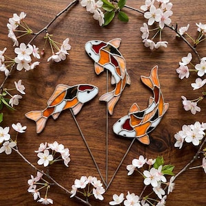 Peut inclure: Trois ornements de poissons koï en vitrail sur tiges métalliques, chacun présentant un motif segmenté de verre blanc, orange et noir. Les poissons sont disposés en groupe, mettant en valeur des lignes de plomb et des couleurs vives.