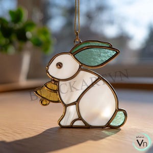 May include: A stained glass bunny ornament with a gold-colored metal frame. The bunny has white, green, and yellow glass panels. The ornament is hanging from a gold-colored chain. The bunny is sitting on a wooden surface.