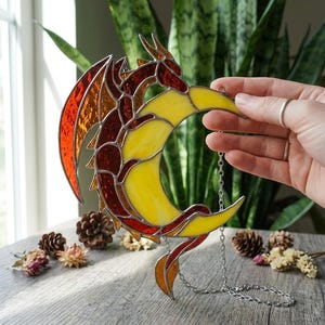 May include: Stained glass dragon and crescent moon suncatcher. The dragon is in shades of red, orange, and brown, with a yellow crescent moon. The suncatcher hangs from a silver chain. Decorative item.