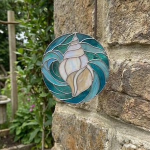 以下が含まれることがあります： 貝殻のデザインが施された丸いステンドグラスのオーナメント。貝殻はクリーム色で、渦巻く青とターコイズのガラスに囲まれ、銀色の縁で縁取られています。金属製のフックから吊り下げられています。