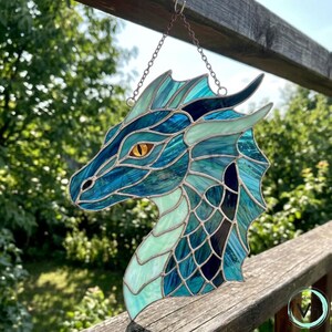 May include: A stained glass dragon head in shades of blue, teal, and black, with a yellow eye. The dragon is suspended from a chain. The artwork is displayed outdoors, with a wooden fence and greenery in the background.
