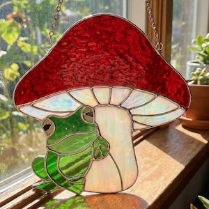 May include: Stained glass mushroom with a red cap and a white underside, featuring a green frog. The artwork is suspended by a chain and is positioned in front of a window, creating colorful light reflections. A whimsical decorative piece.