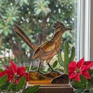 May include: Stained glass roadrunner sculpture with a cactus, set on a wooden base. The bird features shades of brown and amber, with a green cactus. Red poinsettias and greenery frame the artwork.