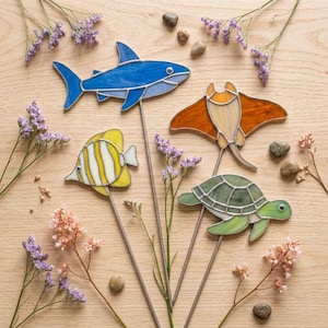 Peut inclure: Quatre créatures marines en vitrail sur tiges métalliques : un requin bleu, un poisson rayé jaune et blanc, une raie orange et une tortue de mer verte. Chaque pièce est assemblée avec des lignes de métal soudées et des fleurs violettes.