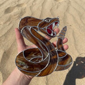 Könnte beinhalten: Ein Buntglas-Suncatcher in Form einer zusammengerollten Klapperschlange mit braun-weiß marmorierten Glassegmenten, silbernen Metalllötlinien, einem roten Glasmund, weißen Glaszähnen und einem segmentierten Rasselschwanz mit zwei kleinen Metallösen.