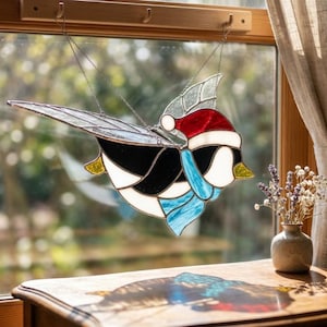 Puede incluir: Un atrapasoles de vidrieras con forma de pájaro que lleva un gorro de Papá Noel rojo y blanco y una bufanda azul. El pájaro presenta segmentos de vidrio negro, blanco y amarillo con alas texturizadas transparentes, suspendido por cadenas metálicas.