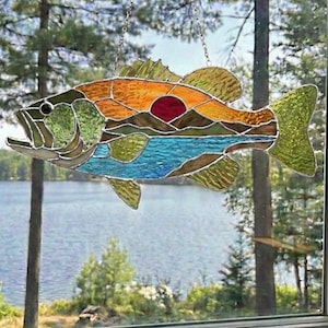May include: Stained glass fish suncatcher with a landscape design. The fish features orange, green, and blue glass, with a red sun and mountain scene. It hangs from a silver chain, ideal for a window decoration.