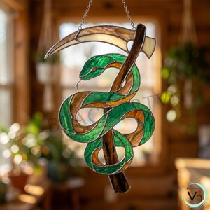 Op de afbeelding: Een glas-in-lood suncatcher met een groene slang die om een bruine zeissteel is gewikkeld. Het zeisblad is gebogen en licht van kleur. Het object hangt aan een zilveren ketting en heeft donkere metalen soldeernaden tussen de glasdelen.