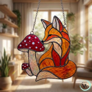 May include: A stained glass suncatcher featuring an orange and red fox sitting beside two red mushrooms with white spots. The fox is constructed from textured glass pieces held together by dark metal solder lines and hangs from a silver metal chain.