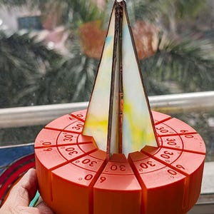 May include: An orange sundial with a triangular stained glass sail. The base is segmented with degree markings, including 30° and 60°. The sail features yellow, blue, and white hues. A unique decorative piece.