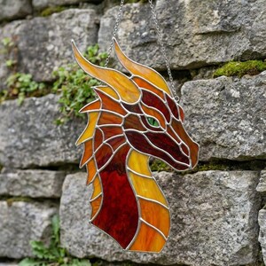 Könnte beinhalten: Glasmalerei eines Drachenkopfes in Rot-, Orange- und Gelbtönen mit grünen Augen. Der Drache hängt an einer Kette. Das Kunstwerk ist vor einer Steinmauer ausgestellt.