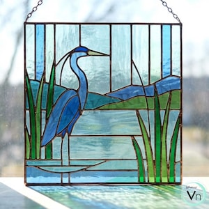 May include: Stained glass panel featuring a blue heron standing in water with green reeds. The artwork uses shades of blue, green, and clear glass, with a chain for hanging. The heron has a long neck and beak, and the background depicts a landscape.