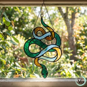 Könnte beinhalten: Bunter Glas-Sonnfänger in Schlangenform mit grünen, blauen und goldenen Abschnitten. Die Schlange hängt an einer Kette vor einem Fenster mit verschwommenem grünem Hintergrund. Der Schlangenkopf befindet sich unten.