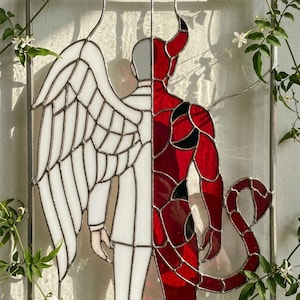 Puede incluir: Vidriera que representa un ángel y un diablo. La mitad del ángel es blanca, con un ala y un traje. La mitad del diablo es roja con detalles negros, cuernos y una cola. La obra está enmarcada con un borde de metal plateado.