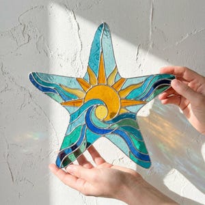 May include: A stained glass starfish suncatcher with a colourful design. The artwork features a yellow sun, blue waves, and a turquoise background. The suncatcher is held up by two hands.