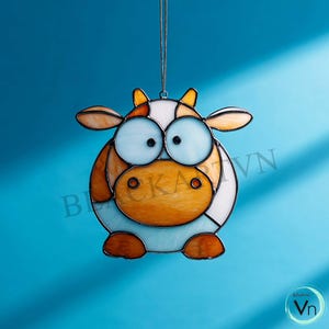 Puede incluir: Un adorno de vaca de vidrieras con un diseño caprichoso. La vaca presenta un cuerpo blanco, detalles en naranja y marrón y ojos azules grandes. Cuelga de una cadena plateada sobre un fondo azul. El adorno es un artículo decorativo.