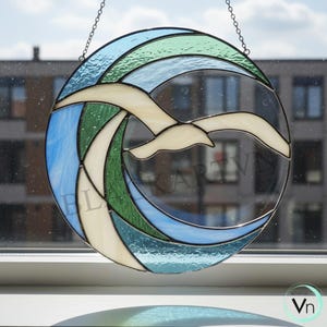 Puede incluir: Una vidriera redonda con un diseño de gaviota en vuelo. La obra de arte presenta una gaviota en vuelo, con piezas de vidrio azul, verde y blanco dispuestas en un patrón de olas. La ventana está suspendida por una cadena.