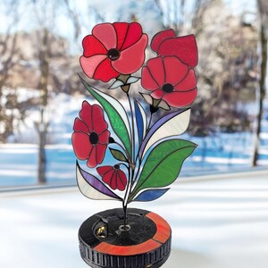Puede incluir: Una escultura floral de vidrieras con amapolas rojas con hojas verdes y azules. La escultura está montada sobre una base negra con un acento rojo. El fondo muestra una escena invernal borrosa.