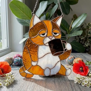 May include: Stained glass cat figurine in shades of orange, brown, and white, holding a black mug. The cat has a sleepy expression and is suspended by a silver chain. A charming decorative piece for cat lovers.