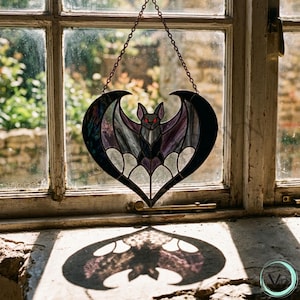 May include: A stained glass bat suncatcher in a heart shape, hanging in front of a window. The bat has purple and black wings, a gray body, and red eyes. The suncatcher casts a bat-shaped shadow.