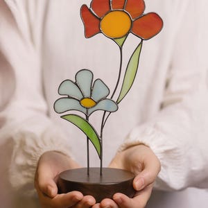 Puede incluir: Una escultura de flores de vidrieras con dos flores estilizadas en tonos naranja, amarillo y azul. Las flores están montadas sobre una base de madera oscura. La escultura está sostenida en las manos de una persona.