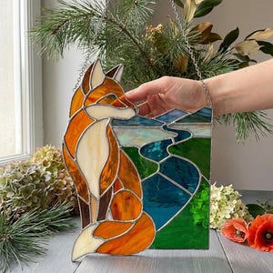 Pode incluir: Vitral com uma raposa em tons de laranja, branco e castanho, num cenário de paisagem azul e verde. A peça é emoldurada com uma corrente prateada, adequada para pendurar. A obra de arte é uma peça decorativa.