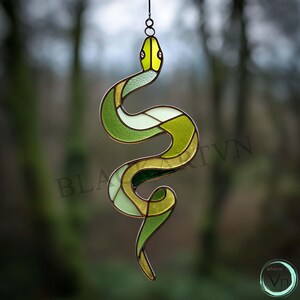 Pode incluir: Um apanhador de sol de vidro manchado em tons de verde e amarelo, com um contorno escuro. A cobra é suspensa por uma corrente de metal, e o design apresenta formas geométricas. O fundo está desfocado, com o texto "BLA TVN" visível.