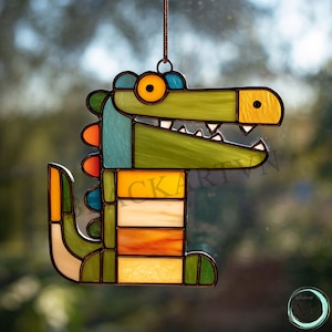 Könnte beinhalten: Bunter Glas-Suncatcher in Alligatorform. Das Design verwendet Grün-, Blau-, Orange- und Gelbtöne. Der Suncatcher wird an einer braunen Schnur aufgehängt und eignet sich zum Aufhängen am Fenster.