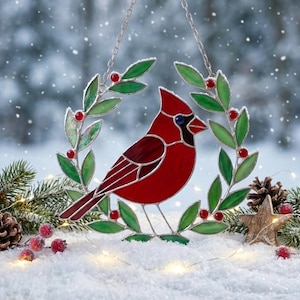 May include: A stained glass suncatcher featuring a vibrant red cardinal bird perched within a circular wreath of green leaves and small red berries. The piece is suspended by a silver metal chain and showcases intricate glass paneling.