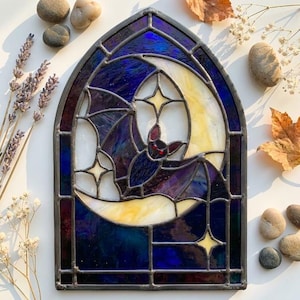 May include: A stained glass panel in a gothic arch shape featuring a dark purple and blue night sky with a large cream-coloured crescent moon. A central bat figure with small red eyes is depicted in flight, surrounded by four-pointed star accents.