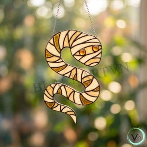 Op de afbeelding: Een glas-in-lood zonnevanger in de vorm van een opgerolde slang met een mummie-achtig verbandpatroon. Het object is gemaakt van gebroken wit en amberkleurig glas, verbonden door donkere metalen soldeerlijnen, met twee ronde amberkleurige glazen ogen.