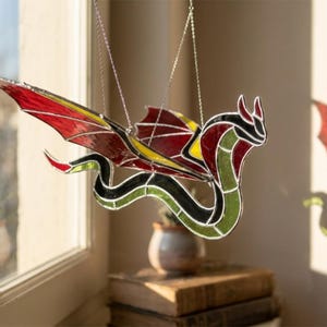 May include: A stained glass dragon suncatcher featuring a long, winding green body with black outlines. The wings are constructed from red and yellow glass panels. The dragon is suspended by thin metal chains, displaying a vibrant colour palette.