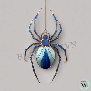 May include: A stained glass spider suncatcher with a copper-coloured frame and hanging chain. The spider features shades of blue and teal glass, with a lighter blue and white abdomen. The legs are long and slender, and the overall design is intricate.