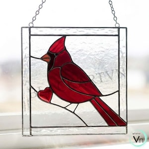 May include: Stained glass window art featuring a vibrant red cardinal bird perched on a branch, holding a heart. The bird is set against a clear background within a square frame, with a chain for hanging. A beautiful piece of home decor.