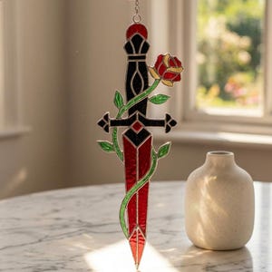 Può includere: Pugnale in vetro colorato con lama rossa, elsa nera e una rosa rossa e gialla. Viti verdi avvolgono il pugnale. Il pezzo decorativo è sospeso da una catena.