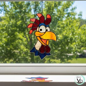 May include: A colorful stained glass rooster suncatcher. The rooster has a yellow beak, red comb, and blue and white accents. It hangs from a chain in front of a window with a blurred green background. A small, colorful object is on the windowsill.