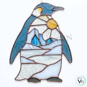 Pode incluir: Apanhador de sol de pinguim em vitral com design de paisagem de inverno. O pinguim apresenta cabeça e asas azuis, com um corpo que mostra uma cena de neve com um sol amarelo. O vidro é delineado com metal cor de cobre.