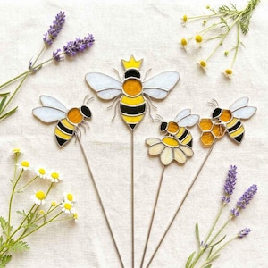May include: Four stained glass bee garden stakes featuring yellow and black striped bodies with white wings. One bee wears a small yellow crown, another rests on a white flower, and a third is positioned next to a yellow honeycomb shape.