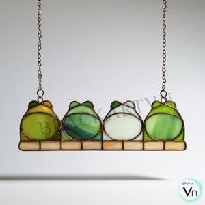 Puede incluir: Colgante de ventana de vidriera con cuatro diseños de ranas verdes. Cada rana tiene un patrón y una variación de color únicos, que van del verde claro al oscuro. Las ranas están conectadas por un marco de metal marrón y suspendidas por cadenas.